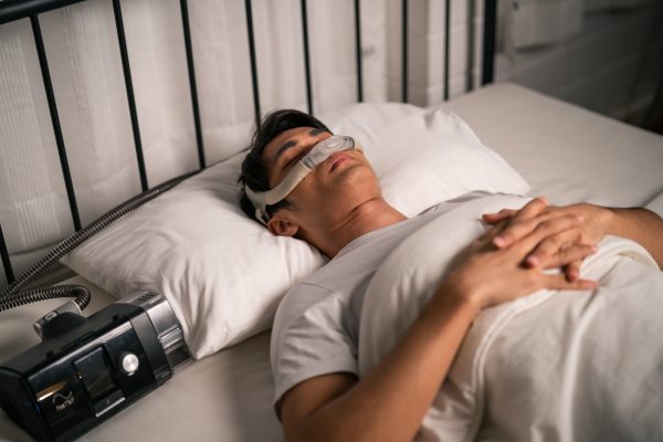 Asian man wearing cpap machine sleeping smoothly all night in bedroom. Attractive handsome healthy male feel happy and relax while lying down on bed, using sleep apnea oxygen mask therapy equipment.