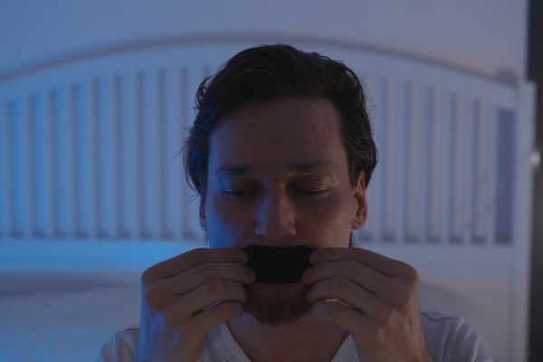 Man in Bed Applying Black Mouth Tape Over Lips with Both Hands Under Dim Blue Light, Anti Snoring and Nose Breathing Concept