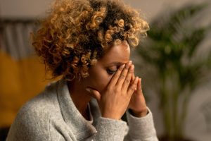 A woman with curly hair in a gray sweater, holding her face, depicting stress indoors